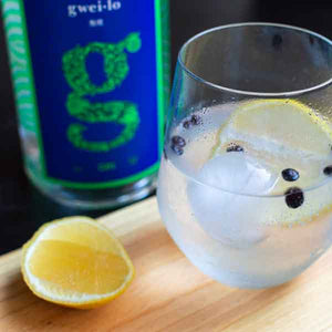 A bottle of Gweilo craft Gin behind a glass of gin and tonic with lemon, juniper berries and ice. This delicious citrus forward gin showcases yuzu and mosaic hops.